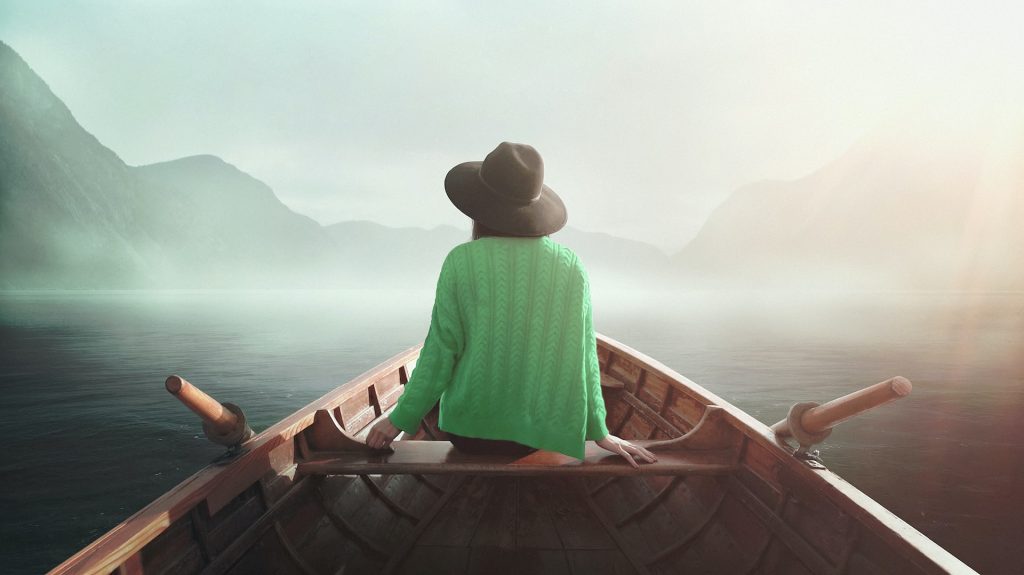 The back view of a lady with a hat on, sited in a boat. The misty view of the mountains ahead and water all around can also be seen.