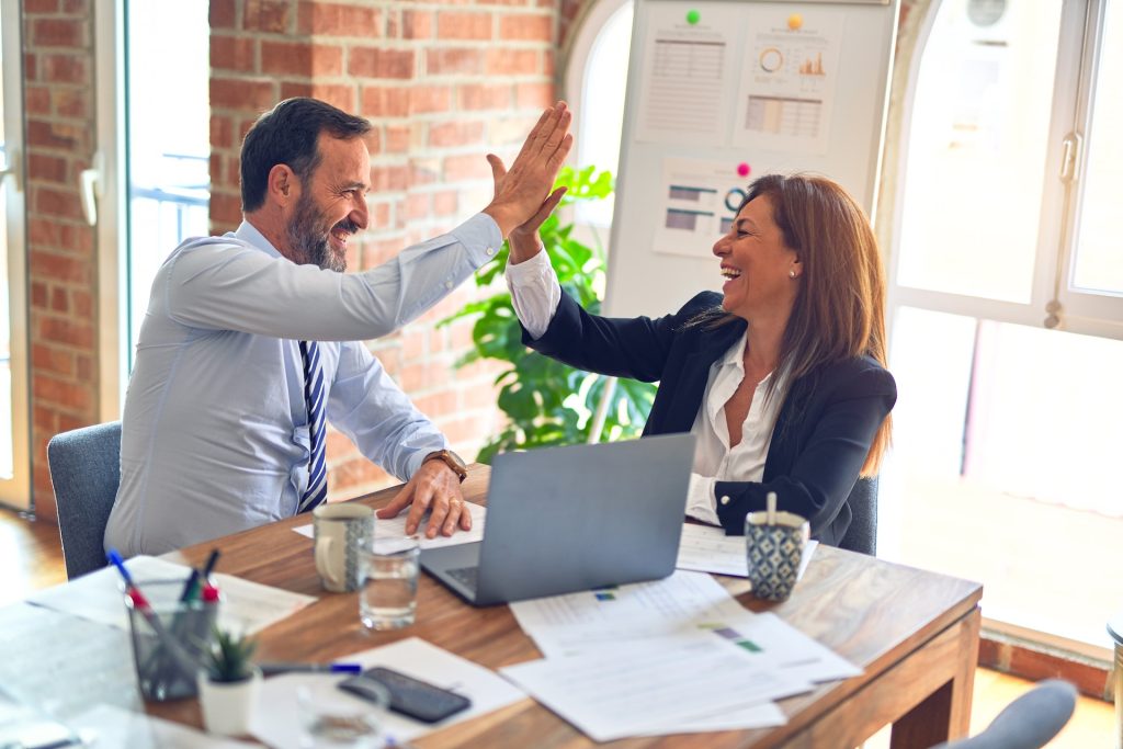 Two people cheerfully sharing a High Five. The laptop, papers, pens, water glass and tea cups on the table with a vision board beside, suggests a goal setting session.