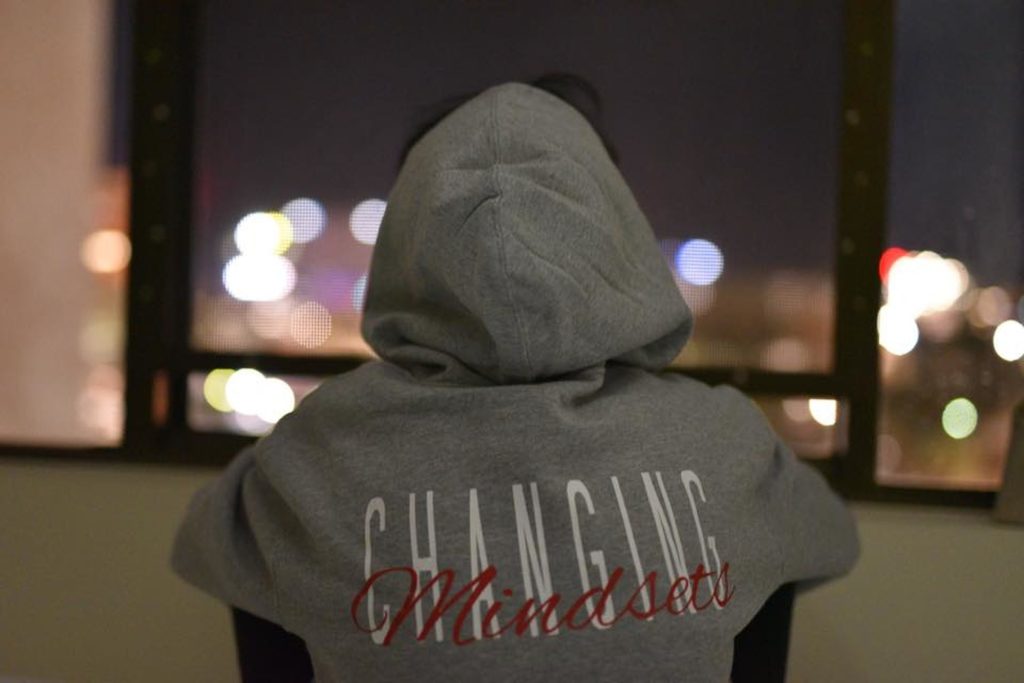 "Changing mindsets" written behind the sweat shirt with head cover of someone sited. He is facing a glass window through which lights are visible. A mindset shift is crucial to overcoming challenges.