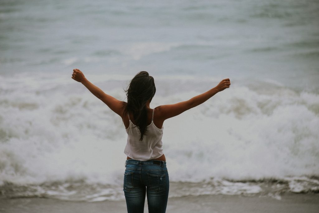 The back view of a lady spreading both hands as water waves approach her at the river banks.