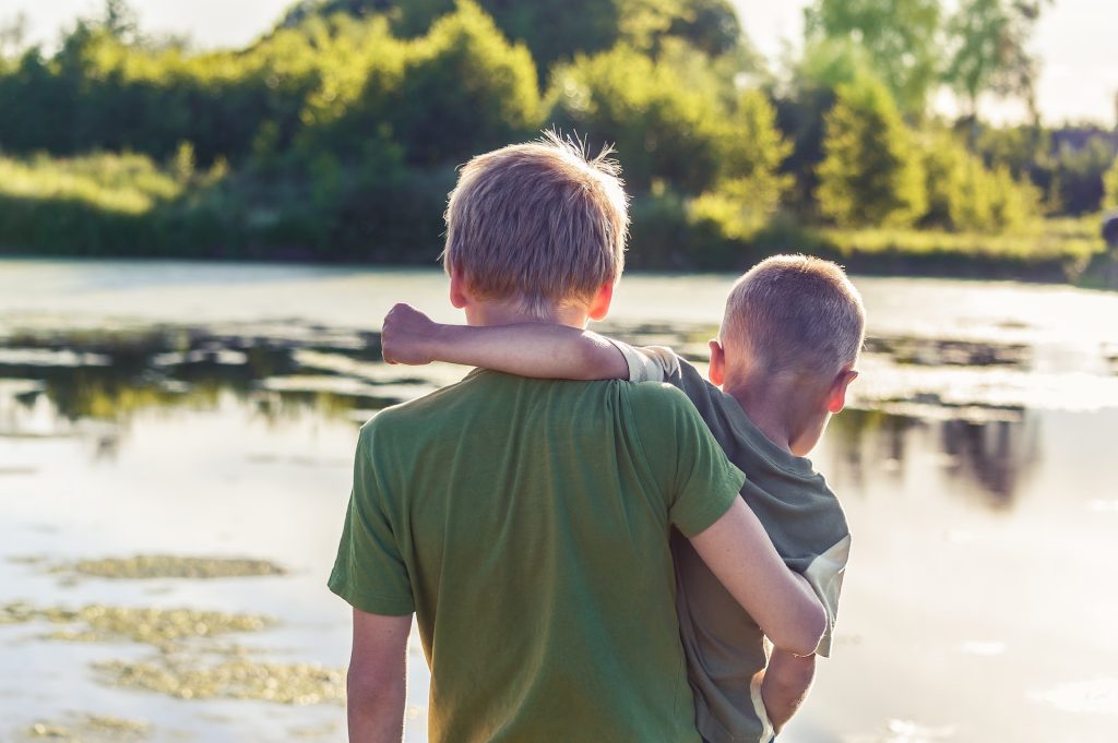 A young boy carrying a younger boy and walking across a water-logged span of land. Overcoming challenges is a catalyst for personal growth and self-discovery.