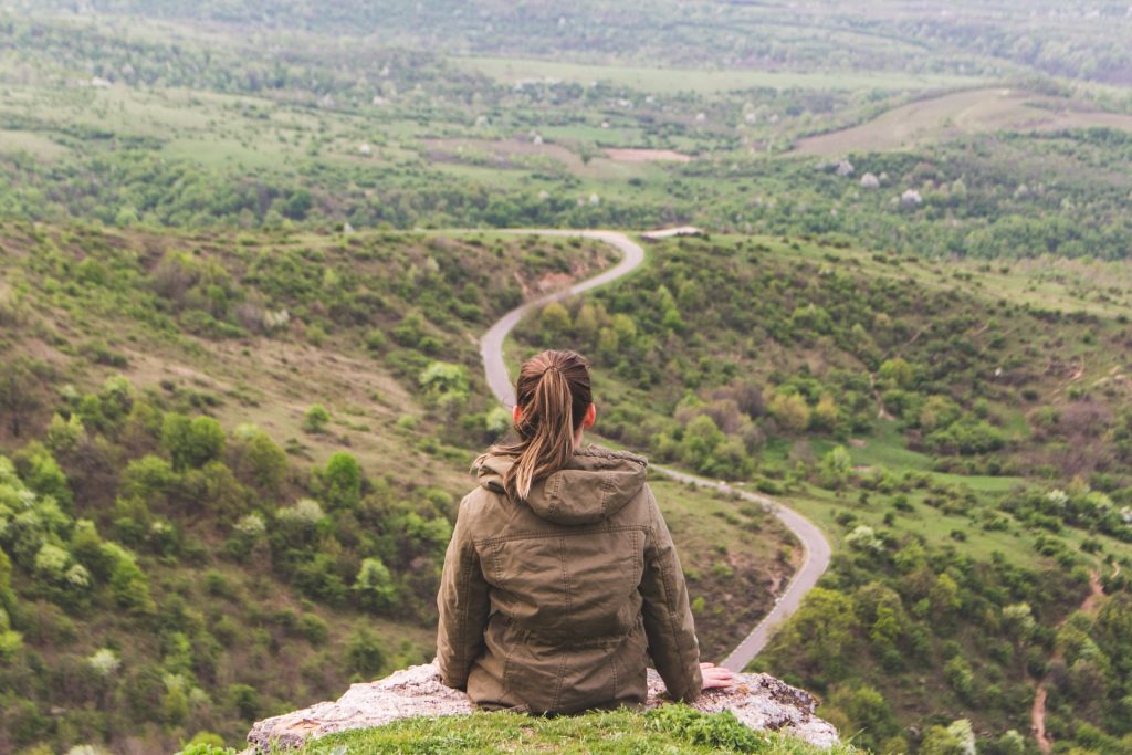 The back of a Lady sitting on a mountain and observing a road path.